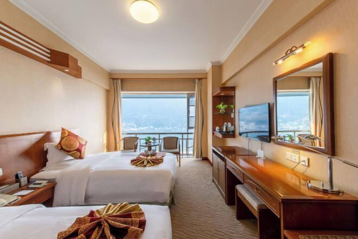A hotel room with two beds, a desk, a TV, and a balcony with a view.