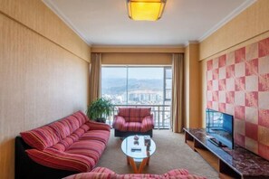 A hotel room with a sofa, armchair, and TV on a stand, with a large window offering a city view.