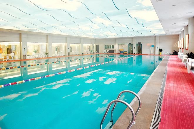 Piscina interna com teto de vidro transparente, assentos à beira da piscina e área com carpete vermelho.