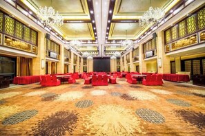 Un amplio salón de conferencias con filas de mesas y sillas, alfombra estampada y un techo alto con candelabros.