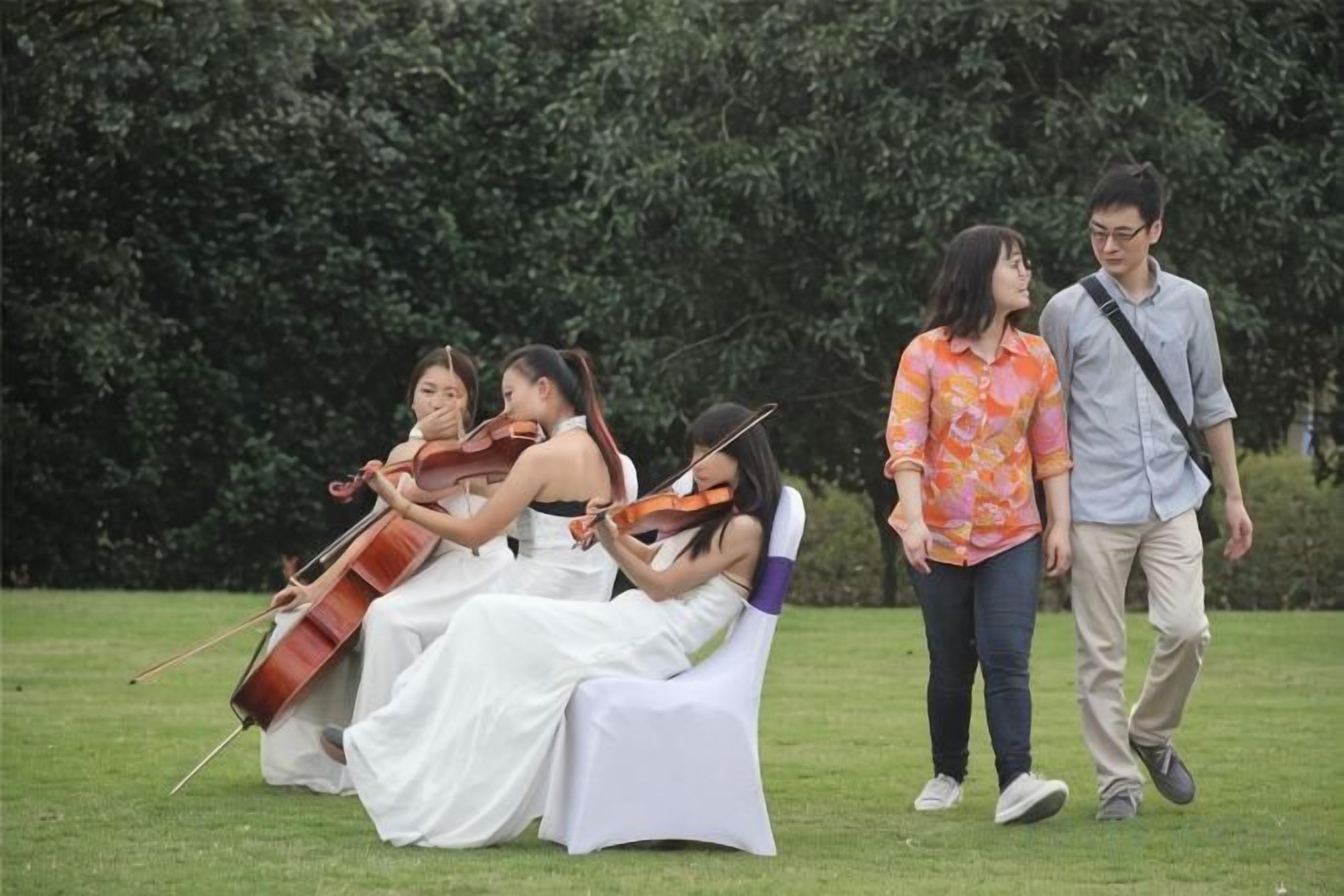 A group of people sitting on white chairs outdoors with greenery in the background.