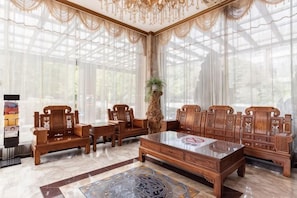 A room with a large wooden table, several wooden chairs, a patterned rug, and a large window with sheer curtains.