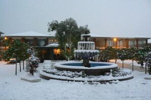 Un espacio exterior cubierto de nieve con una fuente circular, un edificio iluminado y nieve en los árboles.
