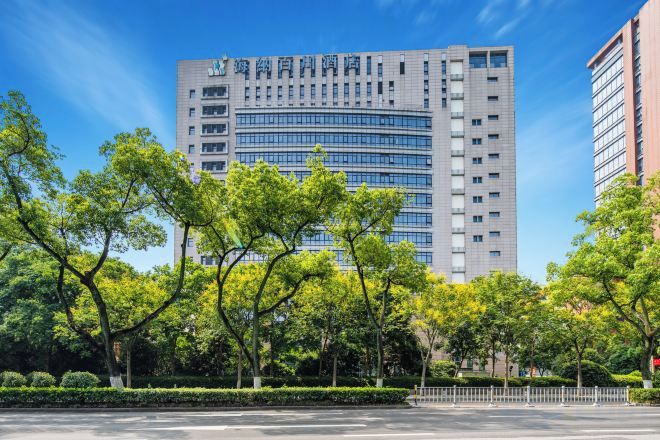 A modern high-rise building with a glass facade, surrounded by well-maintained trees and a clear sky.