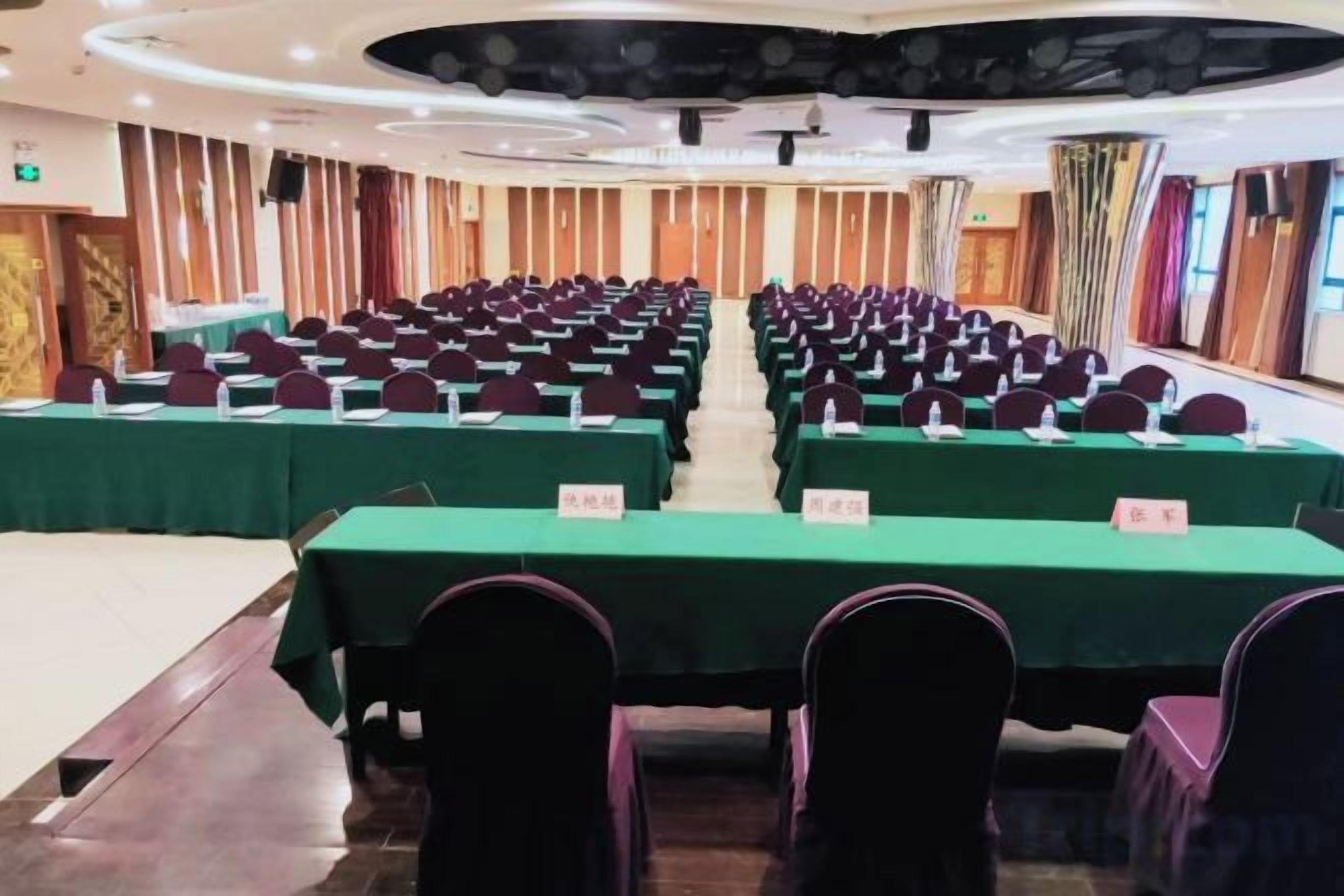 Un salón de conferencias con filas de mesas y sillas, un escenario y un ventanal grande.