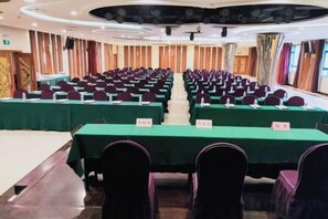 Un salón de conferencias con filas de mesas y sillas, un escenario y un ventanal grande.
