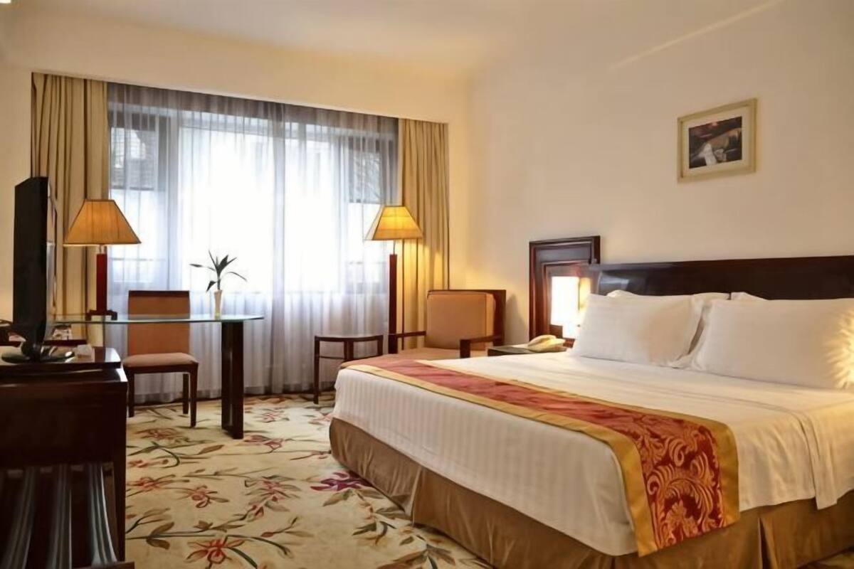 Une chambre d’hôtel avec un grand lit, deux chaises, un bureau, une lampe et une fenêtre avec des rideaux.