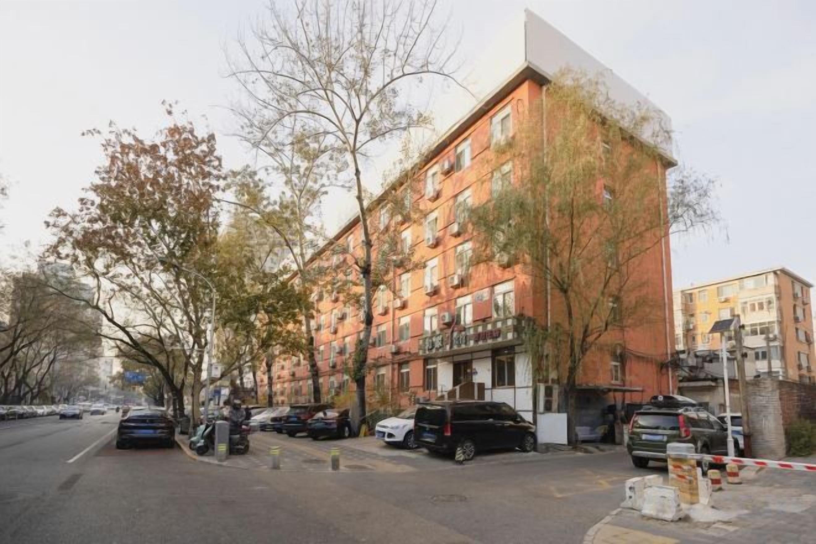 A multi-story building with a brick facade, parked cars, and trees along the street.