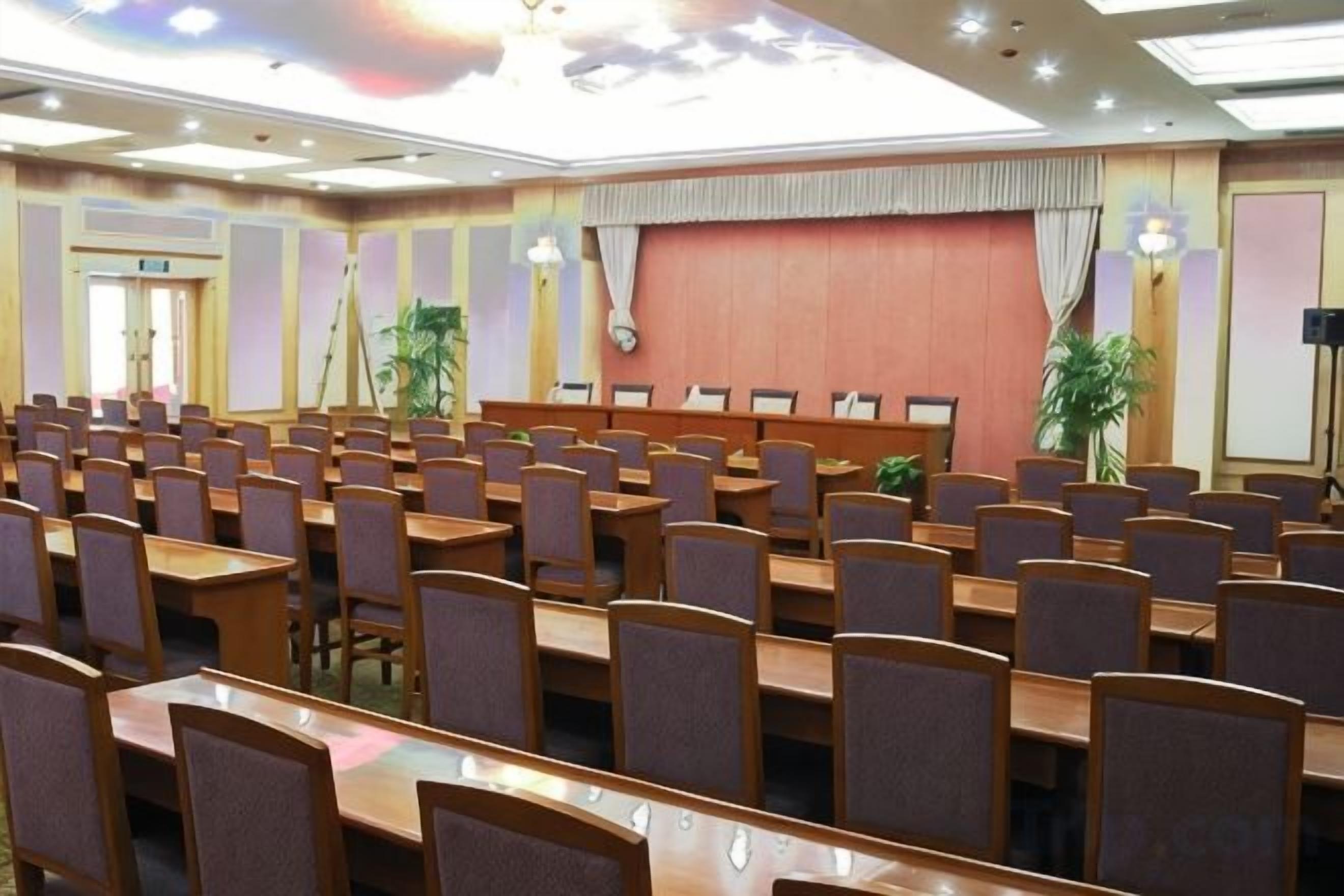A conference room with rows of tables and chairs, a stage with a curtain, and a projector screen.