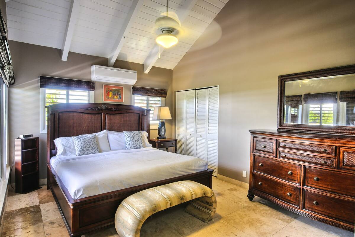 Quarto com uma cama grande, penteadeira de madeira, ventilador de teto e janela com persianas.