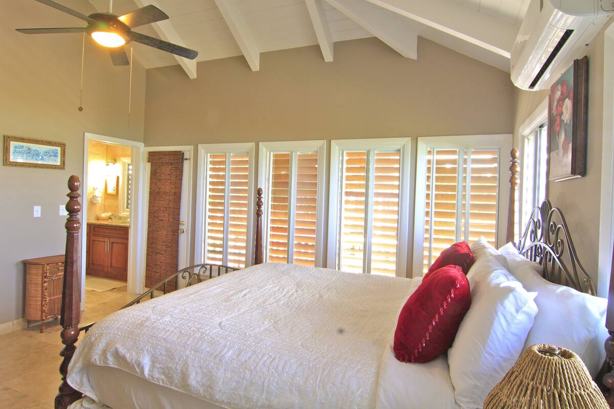 Quarto com cama grande, ventilador no teto e vista para a cozinha através de venezianas abertas.