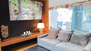 Un soggiorno moderno con una grande TV a schermo piatto, un tavolino consolle in legno con candele, un divano e vista su una piscina e area esterna.