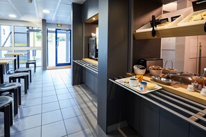 Een moderne ontbijtruimte met een toonbank waarop gebak en drankjes staan, een zithoek met tafels en stoelen, en uitzicht op de omgeving door grote ramen.
