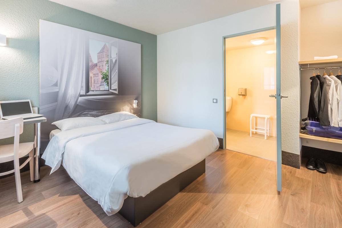 Une chambre d’hôtel moderne avec un grand lit, un bureau avec un ordinateur portable, une armoire et une vue sur un immeuble par la fenêtre.
