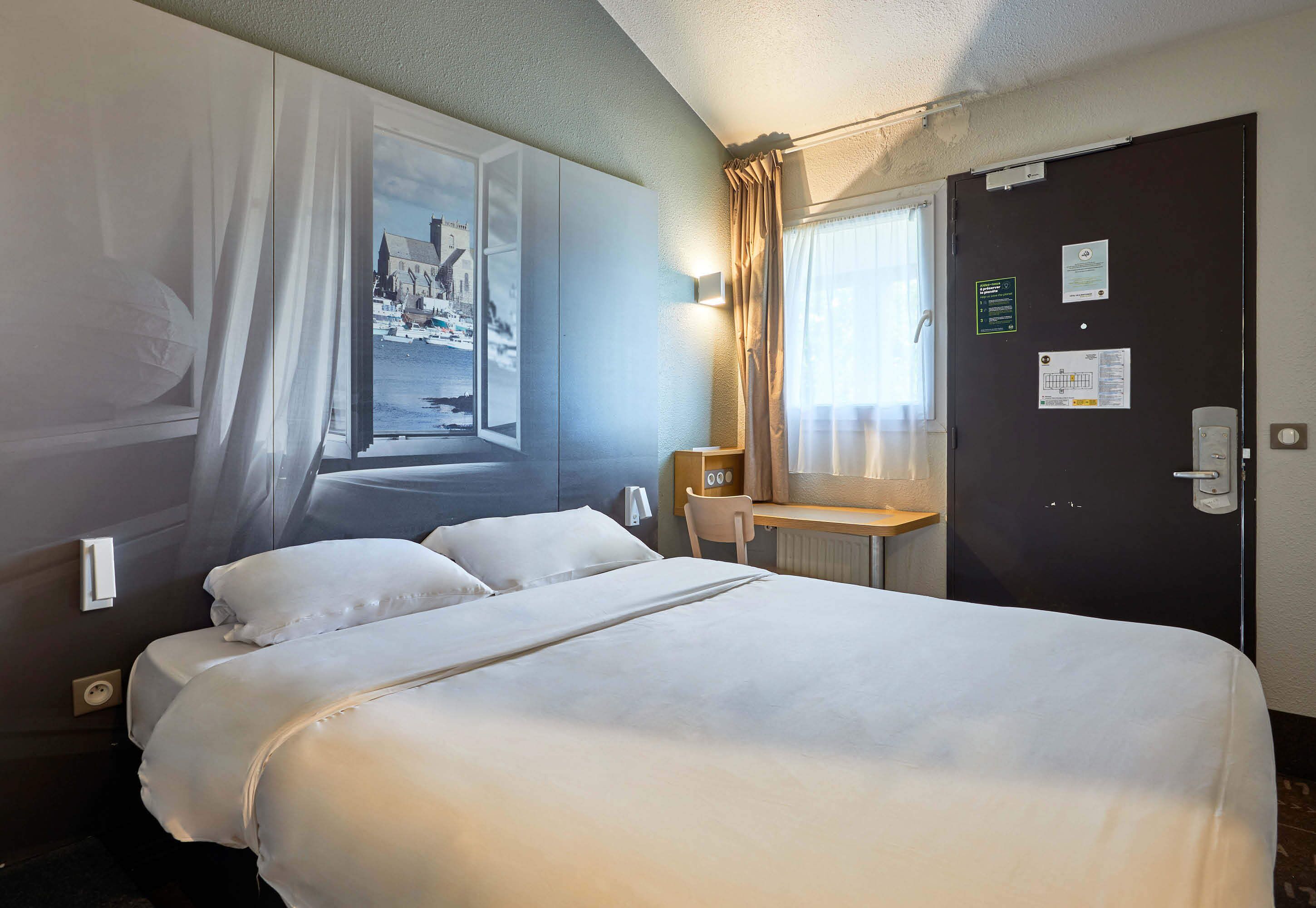 Une chambre d’hôtel comprenant un lit, un bureau, une chaise, une fenêtre avec des rideaux et une porte avec une serrure.