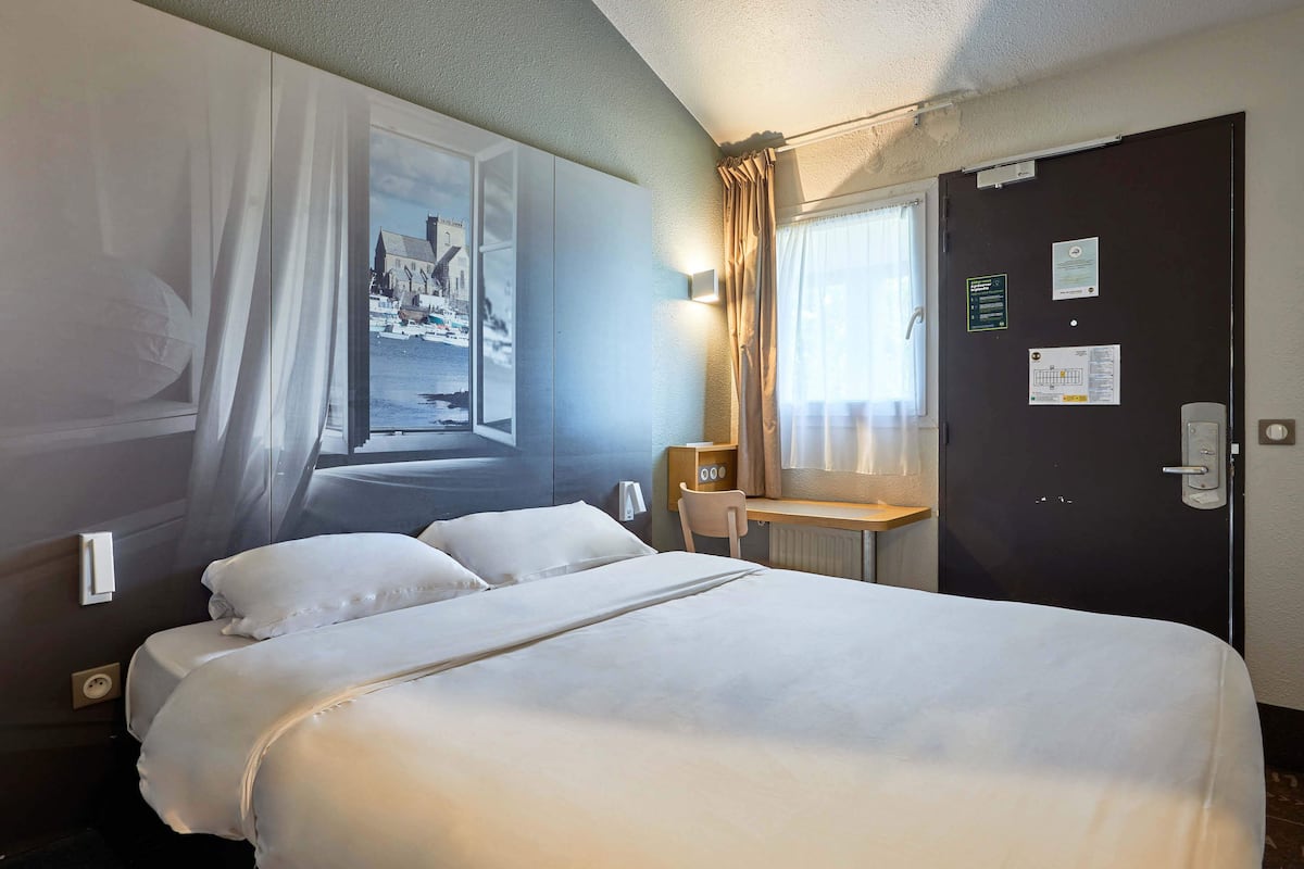 Une chambre d’hôtel comprenant un lit, un bureau, une chaise, une fenêtre avec des rideaux et une porte dotée d’une serrure et d’une pancarte.