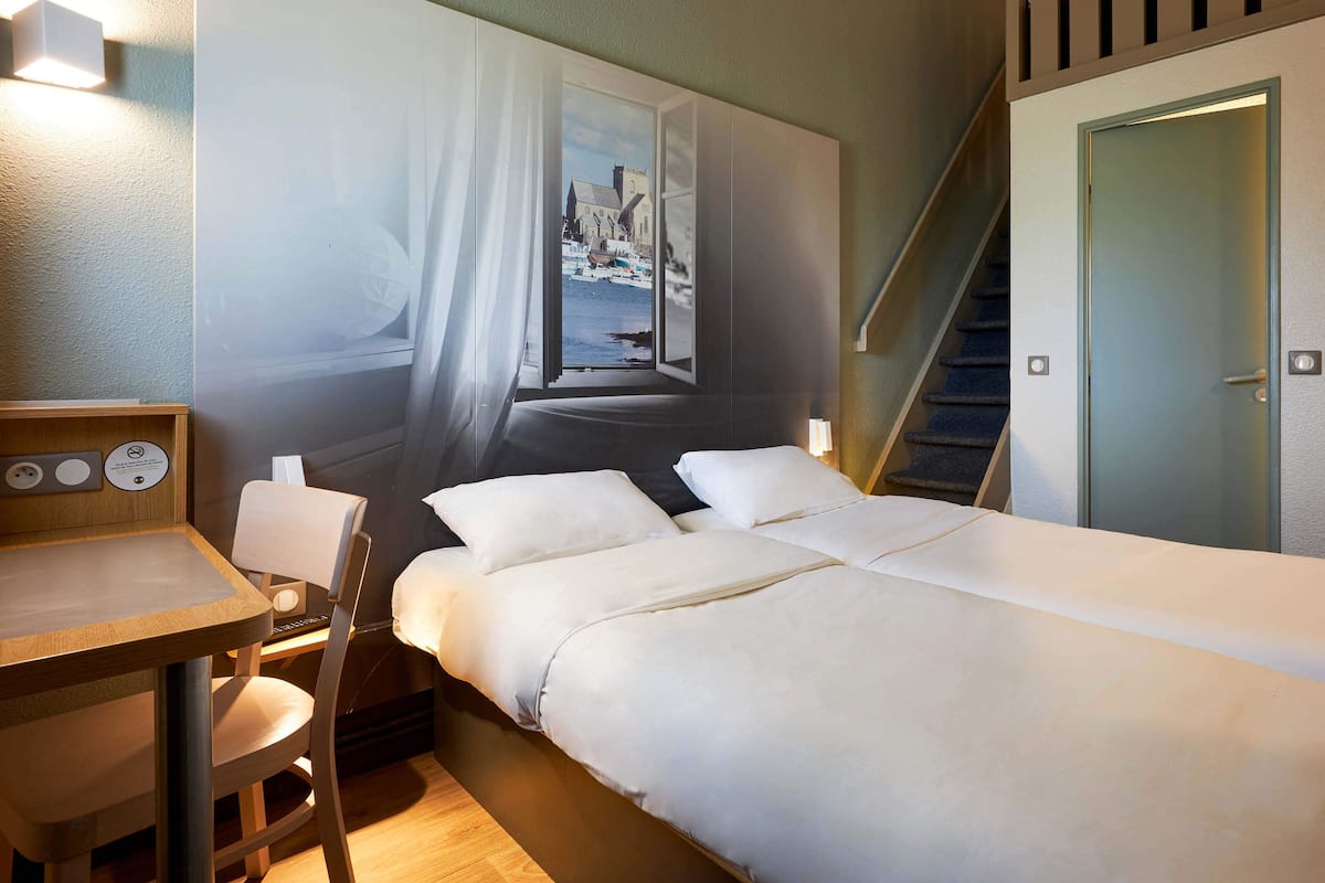 Une chambre d’hôtel comprenant un lit, un bureau, une chaise, une fenêtre donnant sur un immeuble et un escalier.