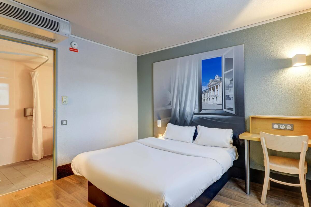 Una habitación de hotel con una cama, un escritorio, una silla, un aire acondicionado y una lámpara en la pared.