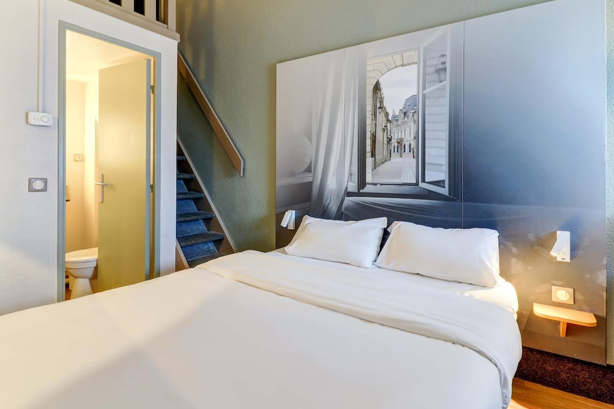 Una habitación de hotel con cama, baño, escalera y un gran ventanal con vista a la calle.