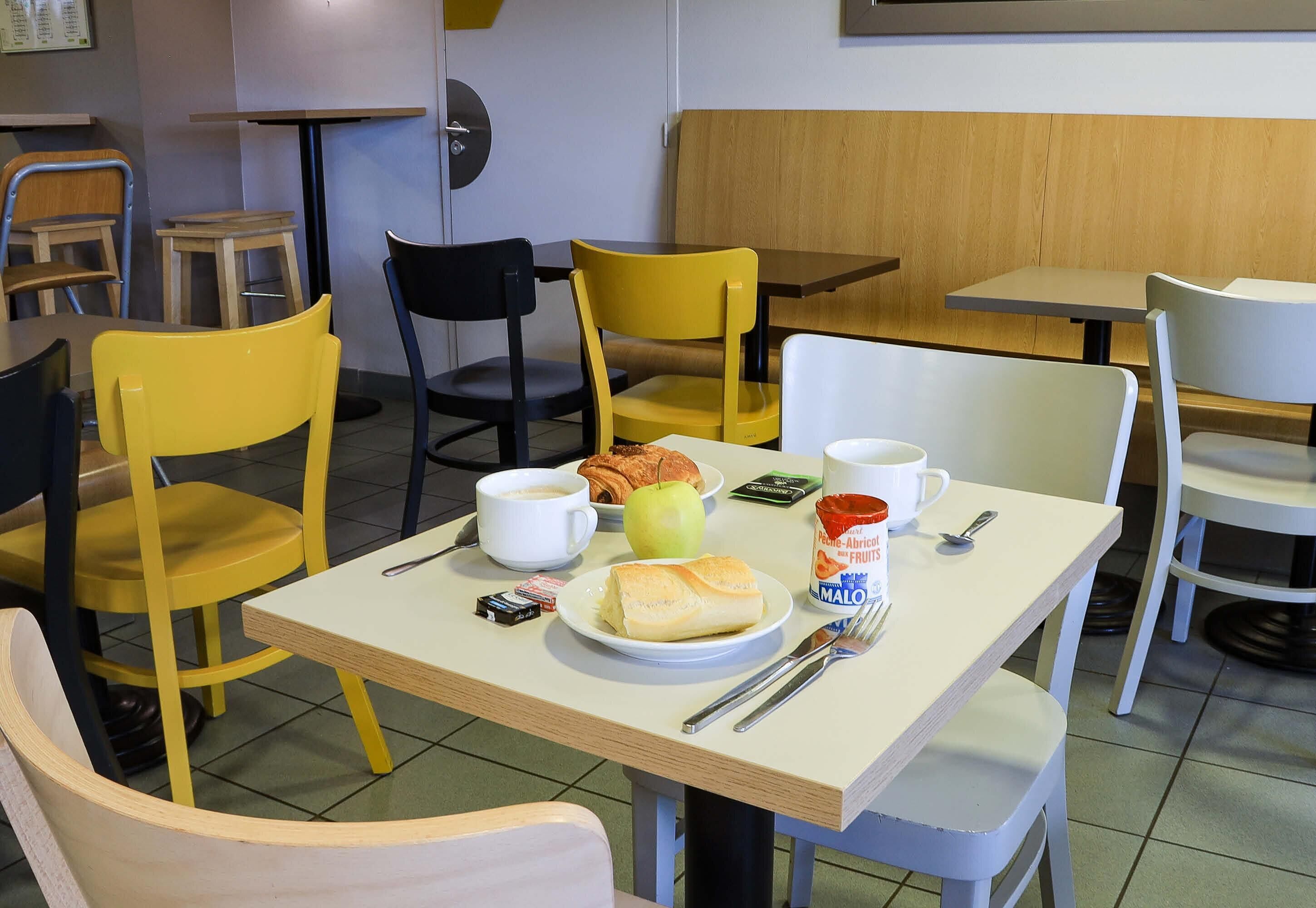 Et spiseområde med borde, stole og en bænk, samt et morgenmåltid med croissant, æble og kaffe.