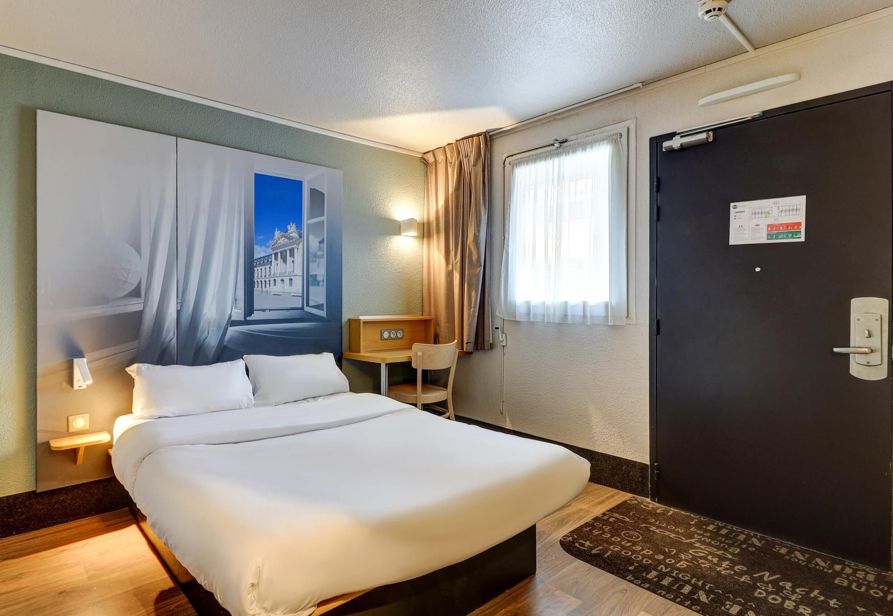 Una habitación de hotel con cama, escritorio, silla, ventana con cortinas y puerta con letrero.