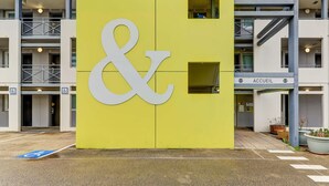 Un edificio amarillo con un gran símbolo de ampersand blanco, un cartel de accesibilidad para sillas de ruedas y un letrero que dice 'ACCUEIL', indicando una zona de recepción.