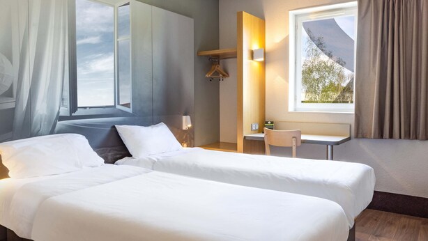 A hotel room with two beds, a desk, a chair, a hanging coat rack, and a window with curtains. - B&B HOTEL DIJON Les Portes du Sud (Acti-Sud) (Marsannay-la-Cote)