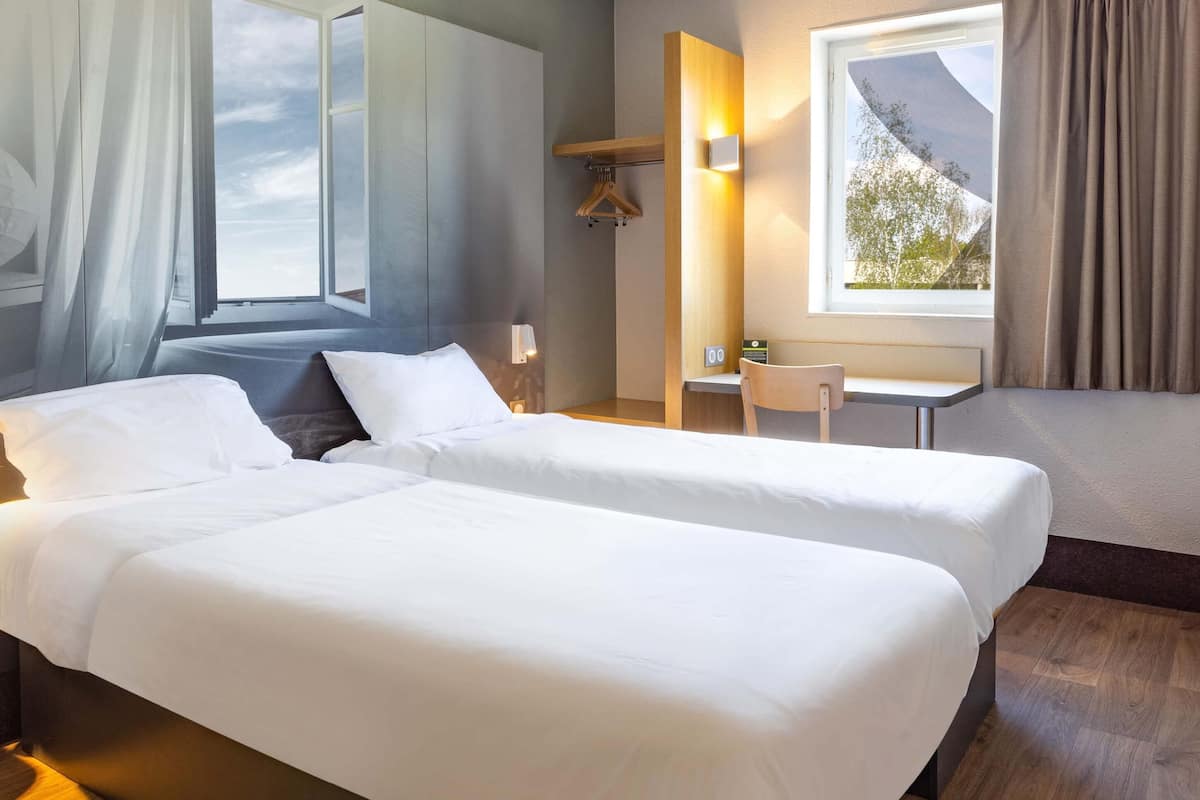 Une chambre d’hôtel avec deux lits, un bureau, une chaise, un porte-manteau et une fenêtre avec des rideaux.