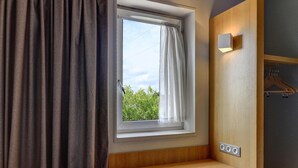 A room with a window, a curtain, a wall-mounted light, a wooden bench, and a wall socket. - B&B HOTEL DIJON Les Portes du Sud (Acti-Sud) (Marsannay-la-Cote)
