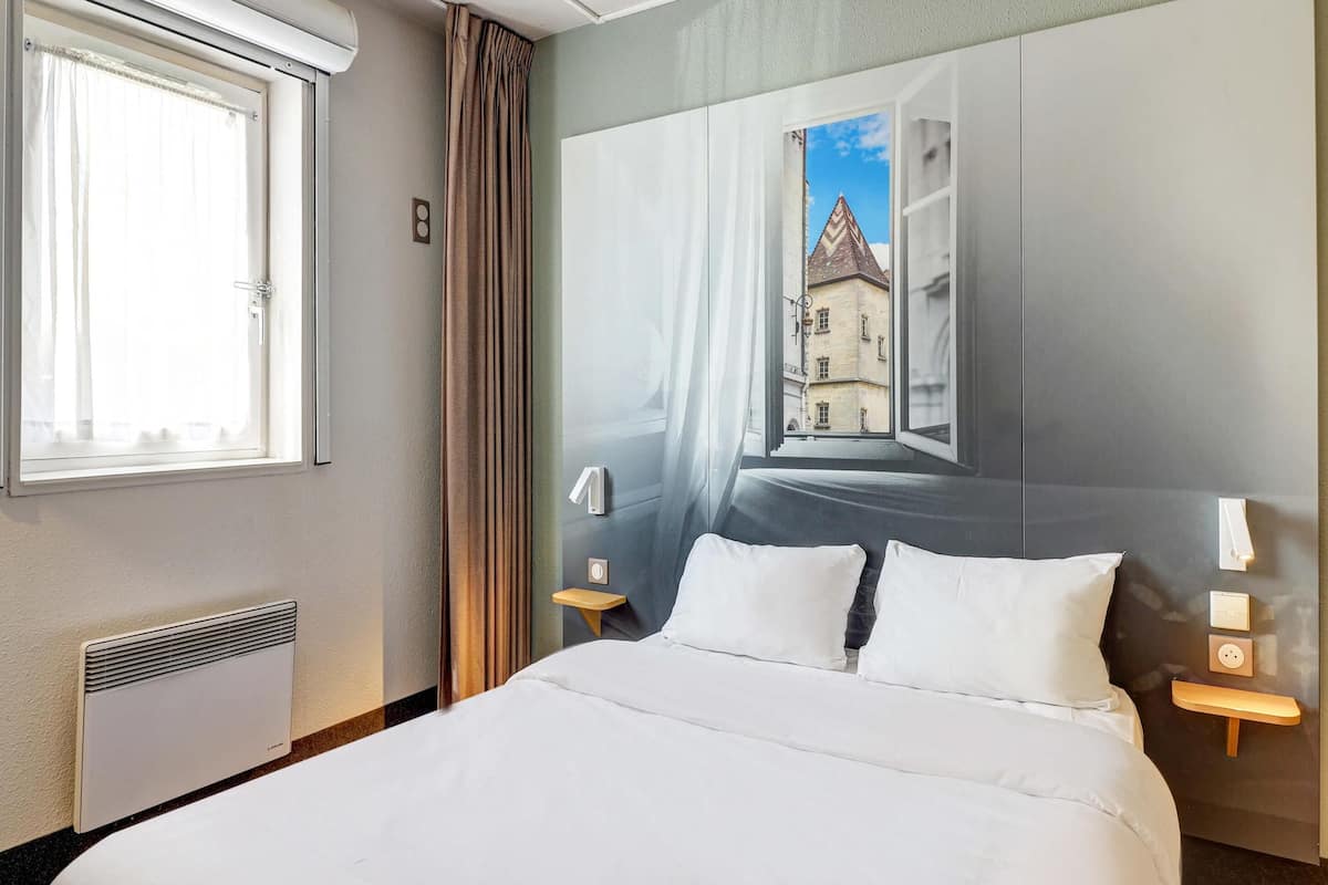 Une chambre d’hôtel avec un lit, une fenêtre avec des rideaux, un radiateur, une lampe fixée au mur et un tableau représentant un bâtiment.