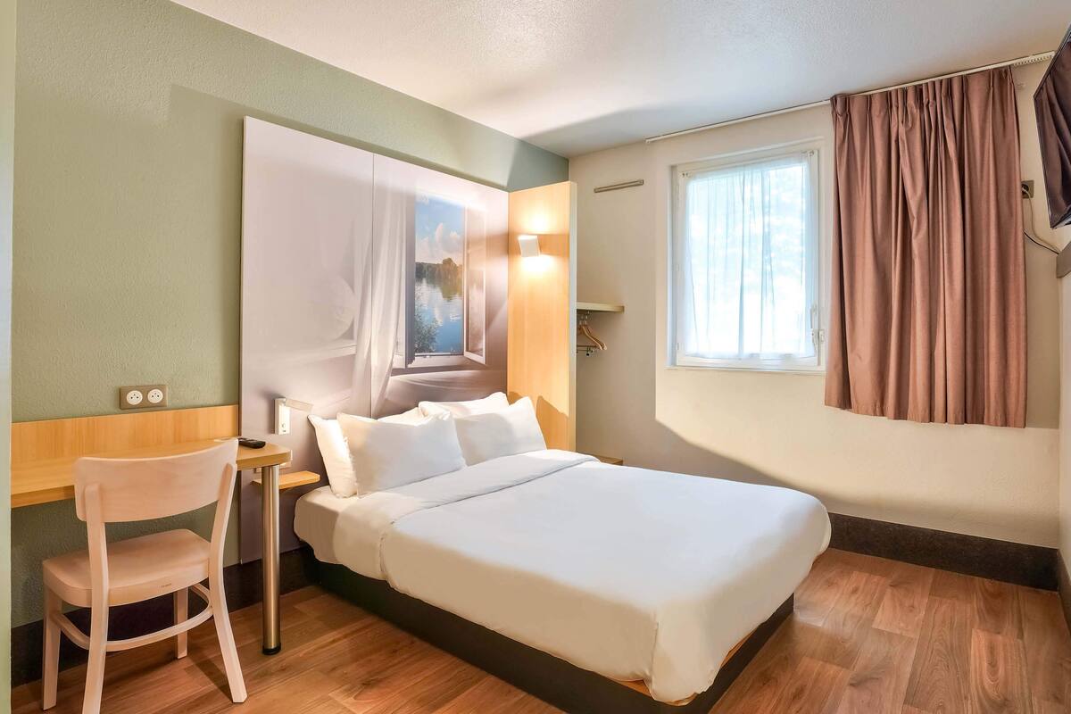 Habitación de hotel con cama, escritorio, silla, un cuadro en la pared, ventana con cortinas y toma de corriente.
