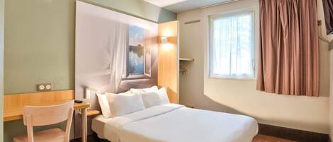Hotelkamer met een bed, bureau, stoel, schilderij aan de muur, raam met gordijnen en een stopcontact.
