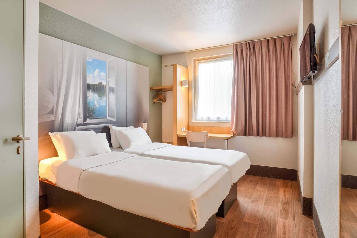 Habitación de hotel con dos camas, un escritorio, una silla, un televisor y una ventana con cortinas.