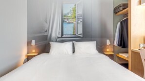A hotel room with a bed, a window with curtains, a view of a bridge and water, and a wooden wardrobe. - B&B HOTEL Goussainville CDG (Goussainville)