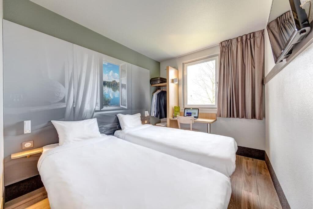 Habitación de hotel con dos camas, un escritorio, una silla, una ventana con cortinas y un televisor montado en la pared.