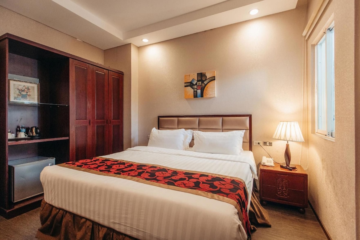 A hotel room with a bed, a nightstand, a lamp, a wardrobe, and a painting on the wall.