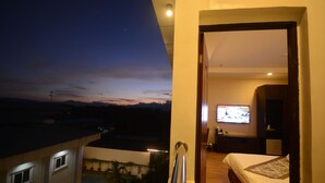 Un balcón con vistas a un atardecer sobre un paisaje, una cama con televisión en la habitación, y una pequeña mesa con una lámpara.