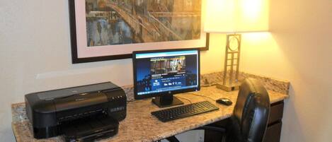 A home office setup with a desktop computer, printer, and a framed cityscape painting on the wall.