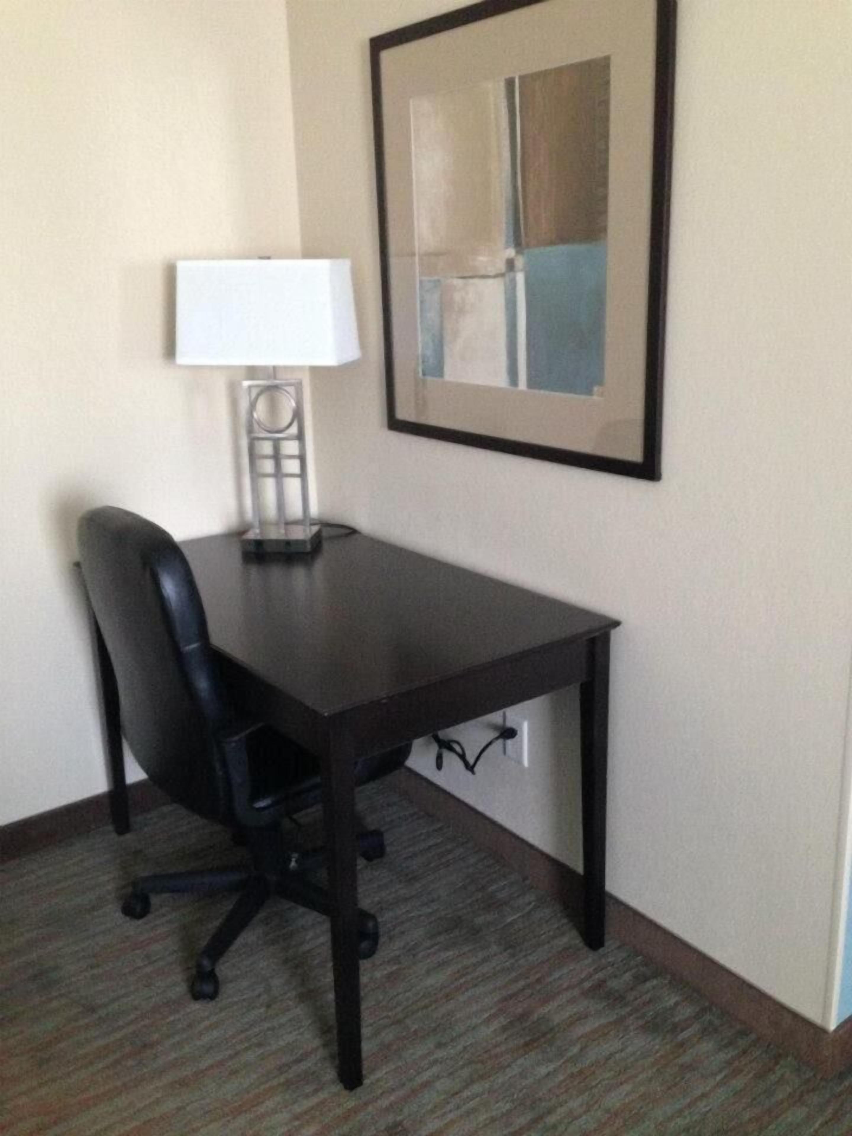 A hotel room with a dark wooden desk, a black office chair, a white desk lamp, and a framed abstract painting on the wall.