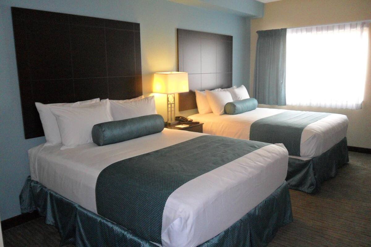 A hotel room with two beds, a nightstand, a lamp, and a window with curtains.