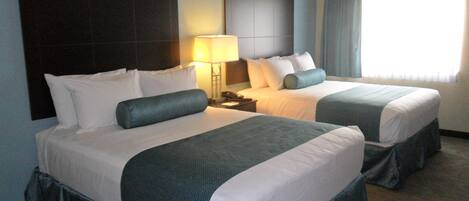 Habitación de hotel con dos camas, una mesita de noche, una lámpara y un ventanal con cortinas.