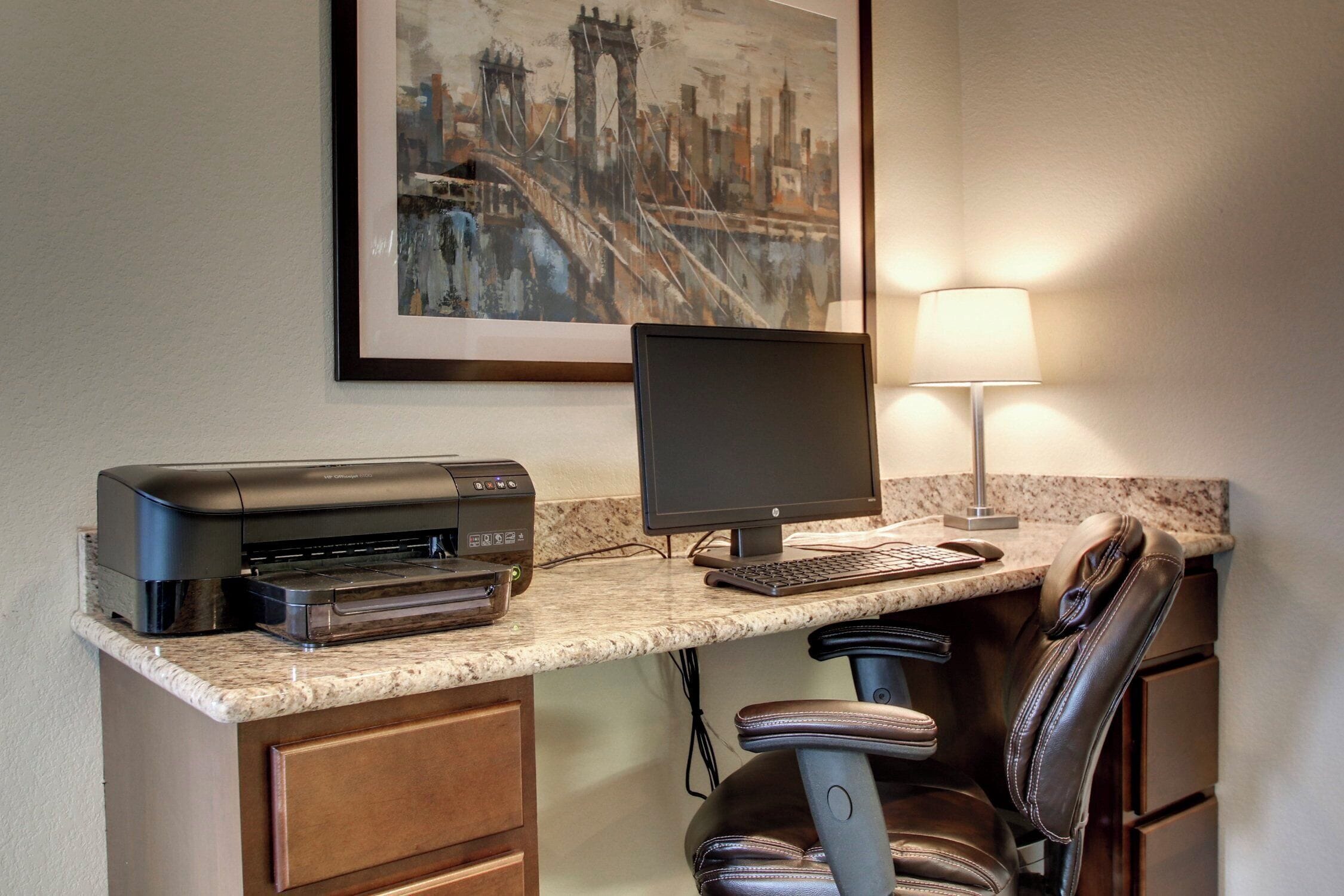 Un espacio de trabajo en un hotel, con computadora de escritorio, impresora y una silla.