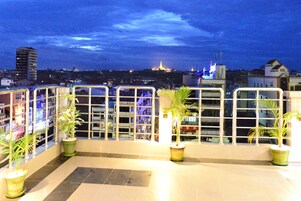 A balcony with potted plants, a tiled floor, and a cityscape view at dusk.