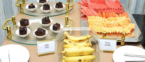 A buffet table with various food items including cakes, bananas, and fruits.