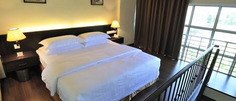 A neatly made bed with white linens, two bedside tables with lamps, and a view of greenery through large windows.