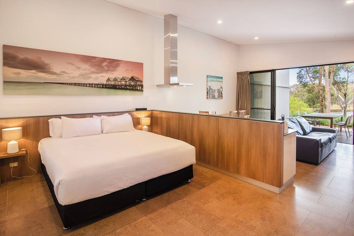 Una habitación de hotel moderna con una cama grande, cabecera de madera, un escritorio pequeño y vistas al exterior a través de un ventanal.