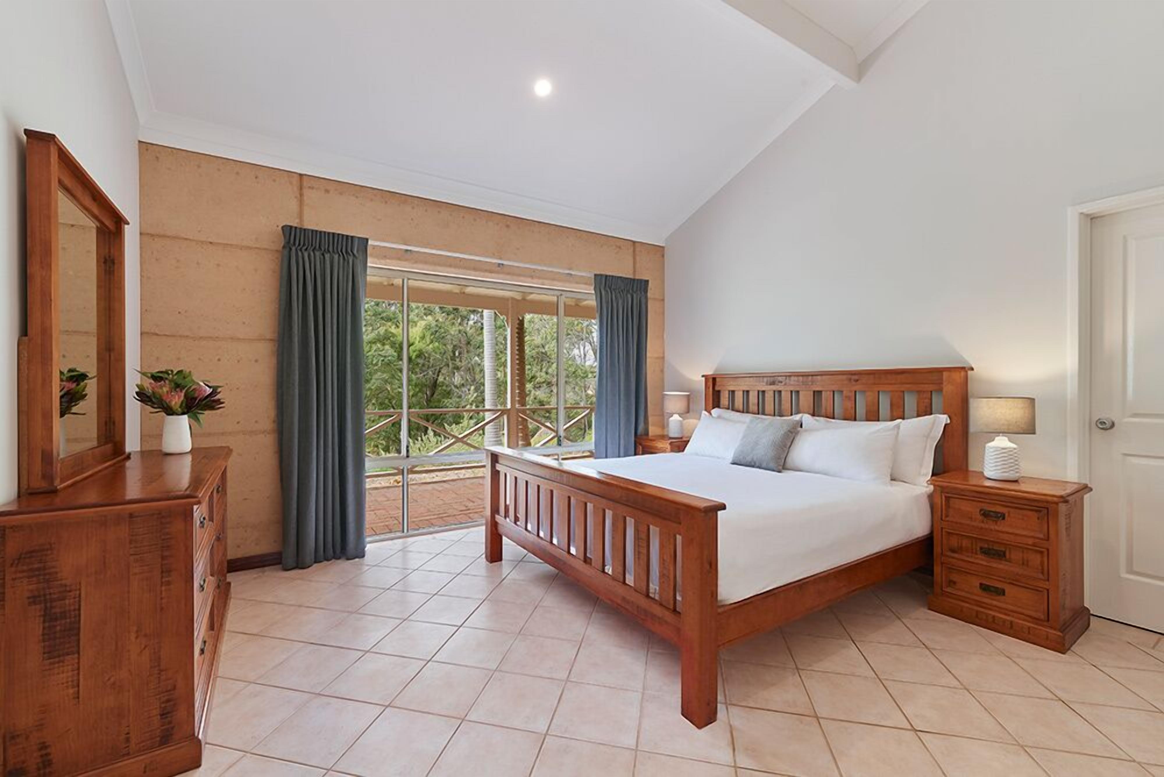 Une chambre à coucher avec un lit en bois, une table de chevet, une commode et une grande fenêtre avec des rideaux.