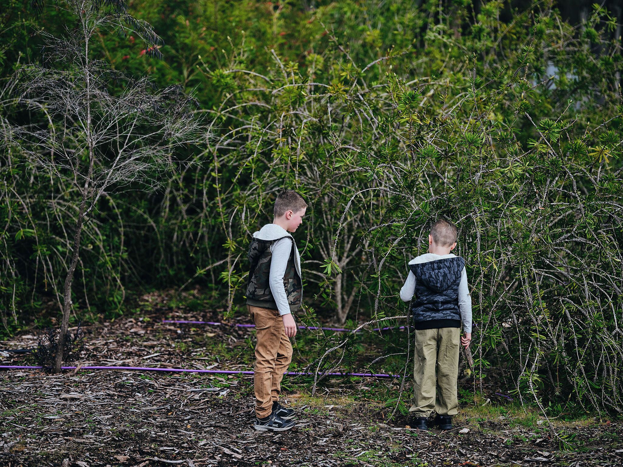 Two children standing in a garden with dense foliage and a purple hose in the background.