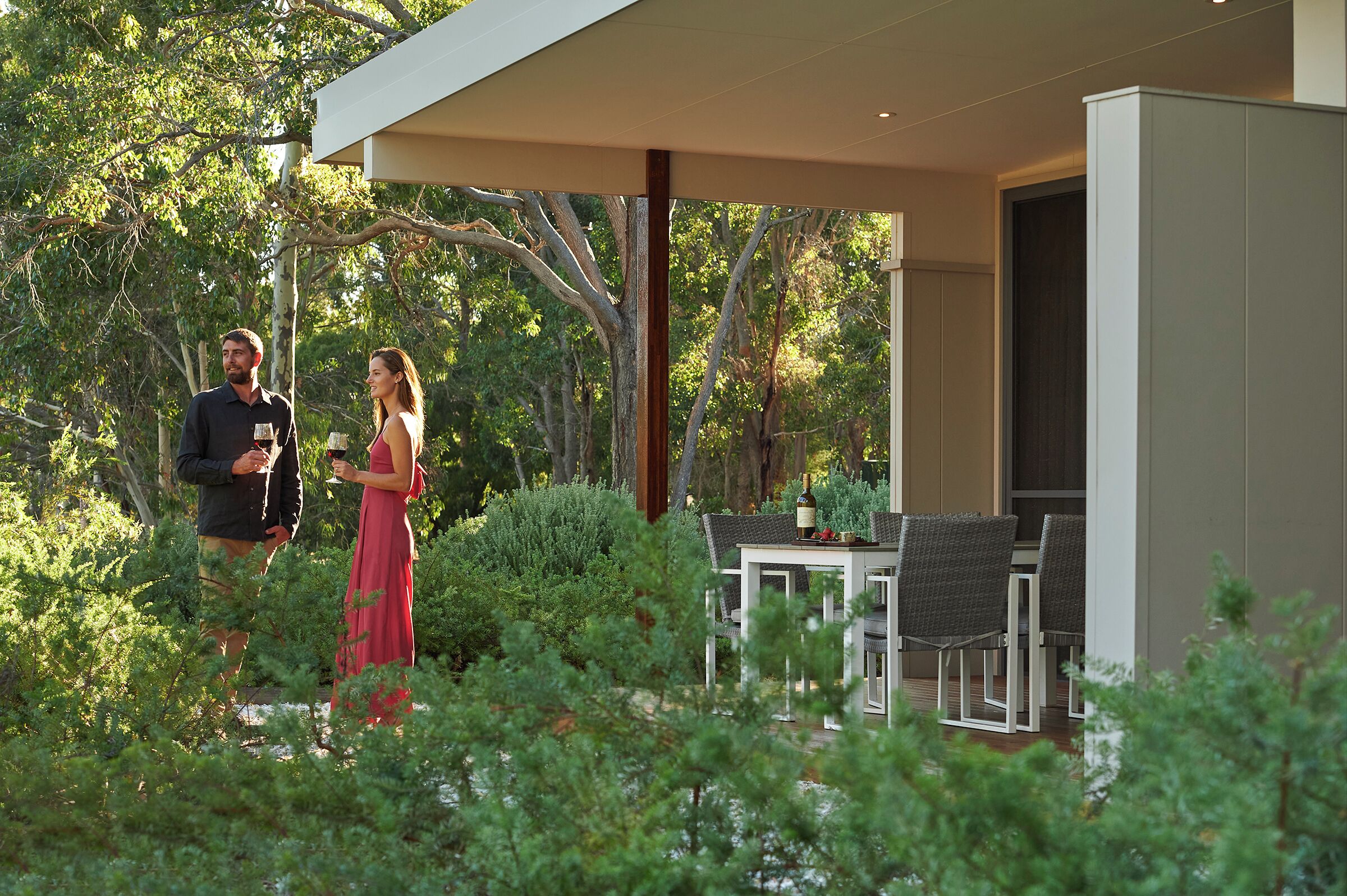 A covered outdoor seating area with a table and chairs, surrounded by lush greenery.