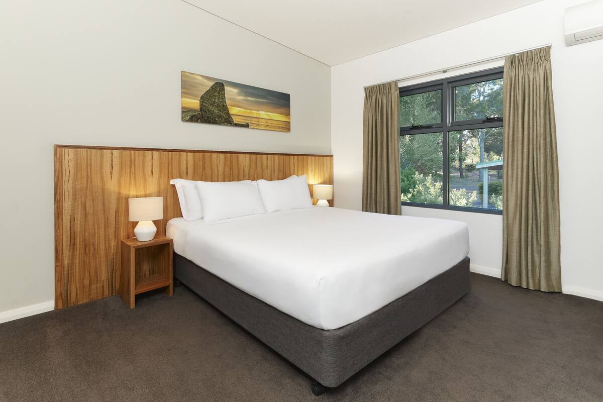 Una cama bien tendida con un cabecero de madera, una mesita de noche con una lámpara y una ventana con cortinas.